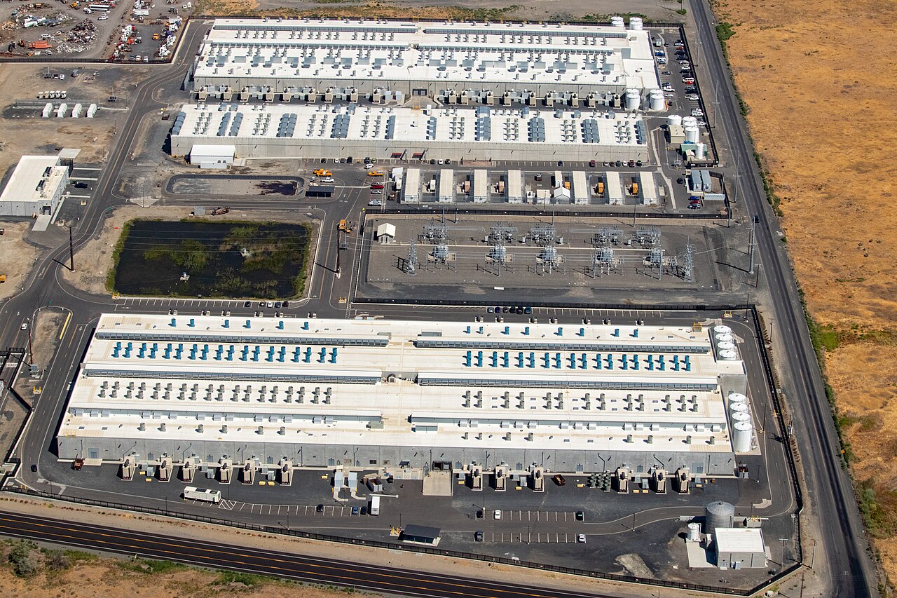 Aerial view of a large-scale Amazon Web Services (AWS) data center complex in Oregon, showing white-roofed buildings, cooling units, electrical substations, and surrounding infrastructure.